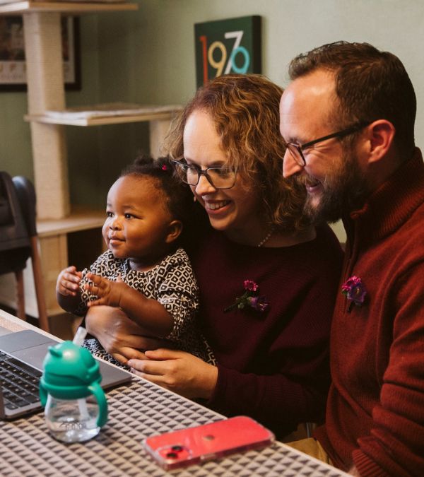 Liz, her husband, Jarrett, and their baby daughter sit at the table facing a computer screen as if in a happy video call.