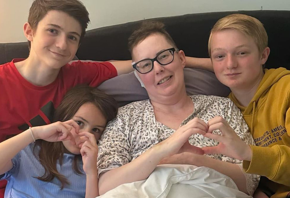 Julie Rock rests and recovers after breast cancer treatment with three of her young children gathered around her making the heart symbol with their hands.
