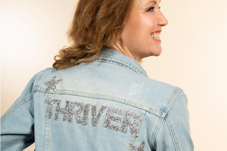 A white woman shows the back of her denim jacket. It says "Thriver" in large, sparkly capital letters.