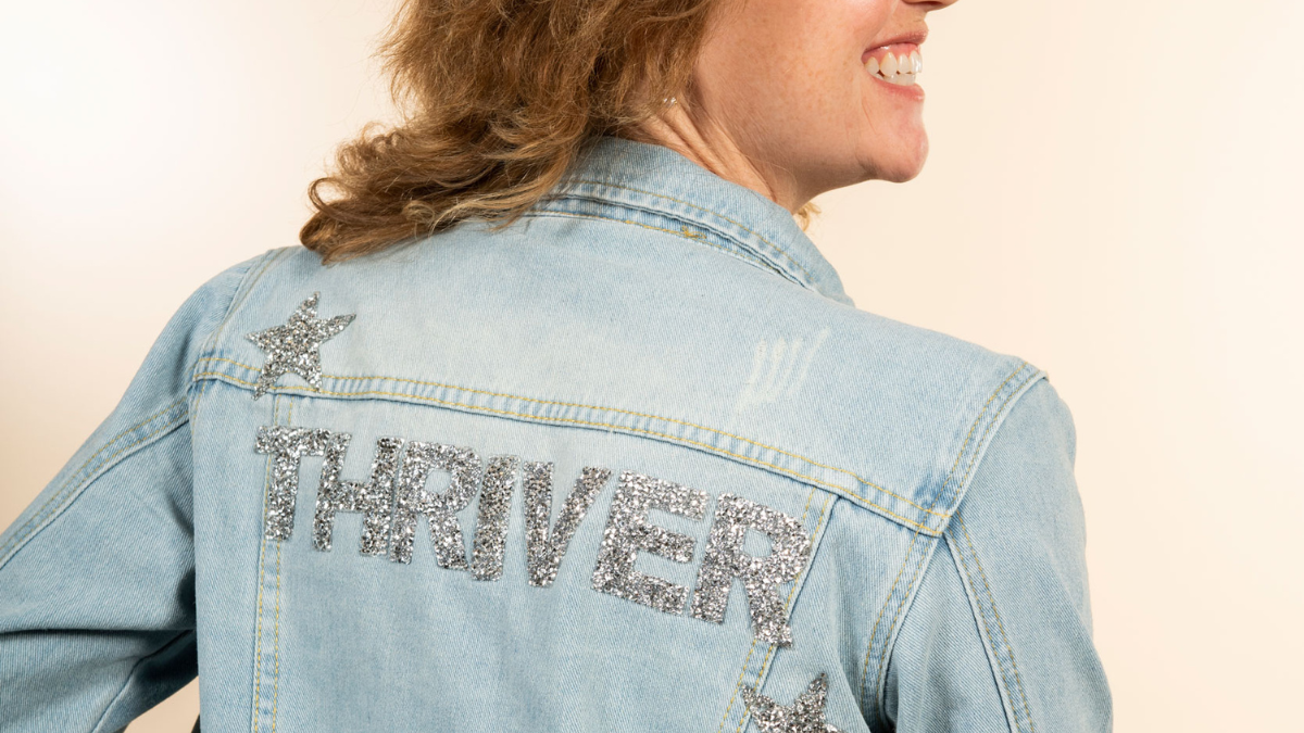 A white woman shows the back of her denim jacket. It says "Thriver" in large, sparkly capital letters.
