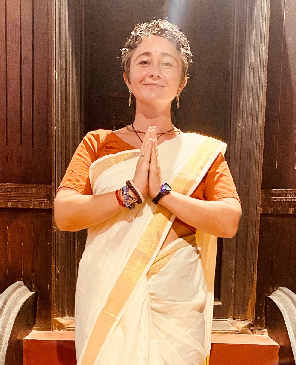 Wearing a sari, Jenna Antonelli clasps her hands in grateful prayer in front of a dark wood temple.