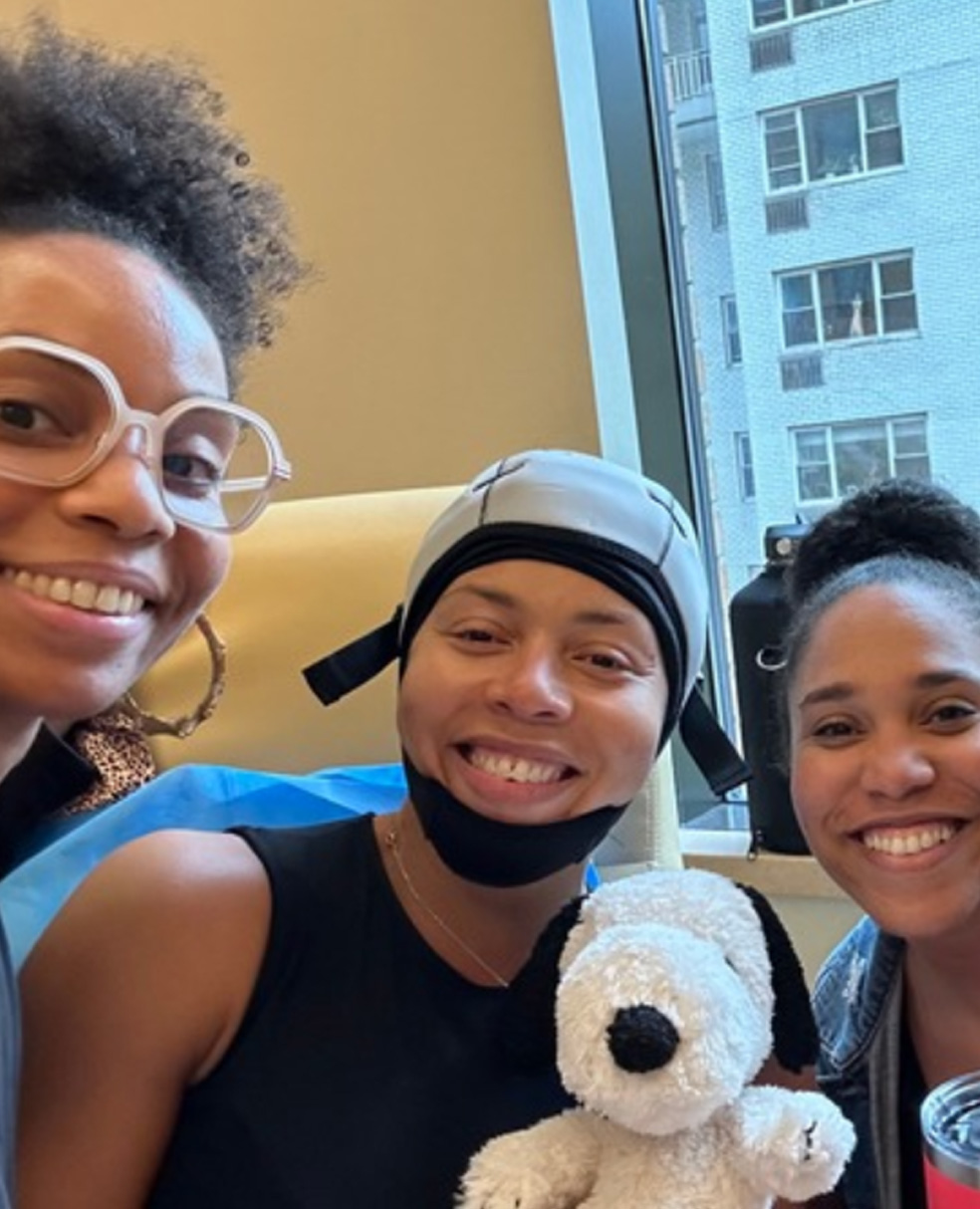 Kia Lee smiling in a chemo chair with a scalp cooling cap on and three family members are smiling around her.