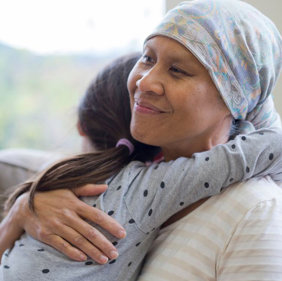 Seated in an armchair, a woman, who is wearing a headscarf and impacted by cancer, is hugged meaningfully by her granddaughter.