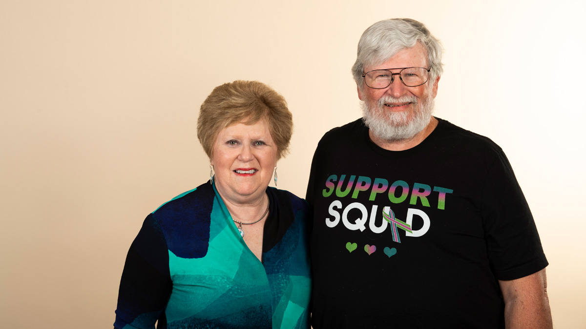 An older white couple smiling. The man's shirt reads, "Support squad."