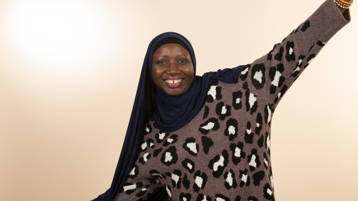 A Black woman wearing a hijab and a cheetah-print sweater, smiling with her arms outstretched.