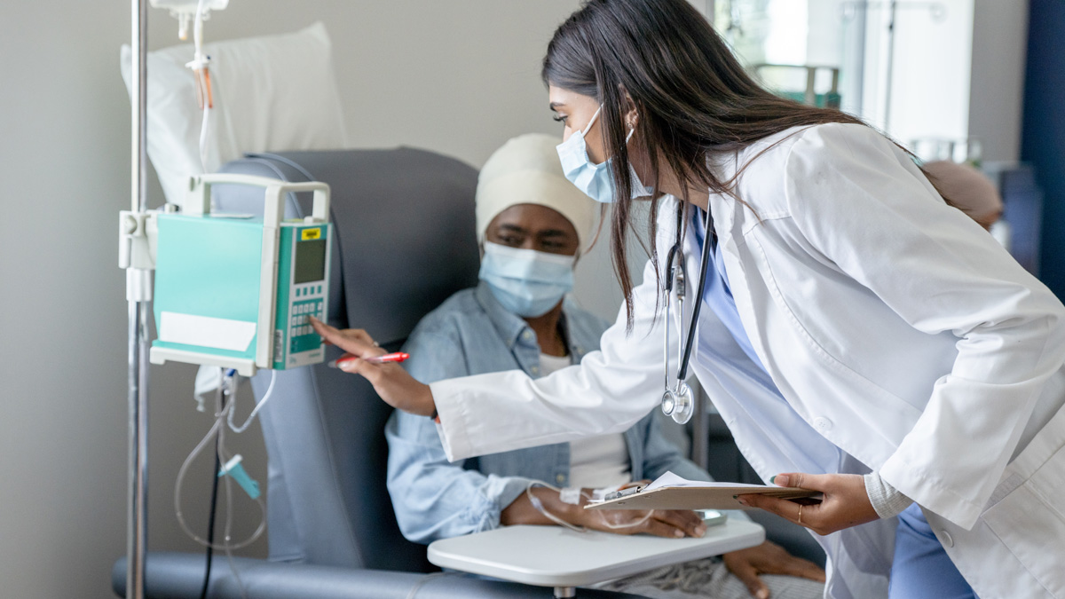 A Black woman sitting in a chemo chair, while an Indian doctor presses buttons on the infusion machine