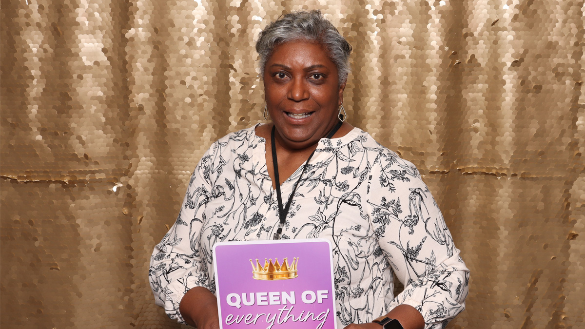 An older Black woman smiling in front of a gold, sparkly backdrop, while holding a sign that reads "Queen of everything"