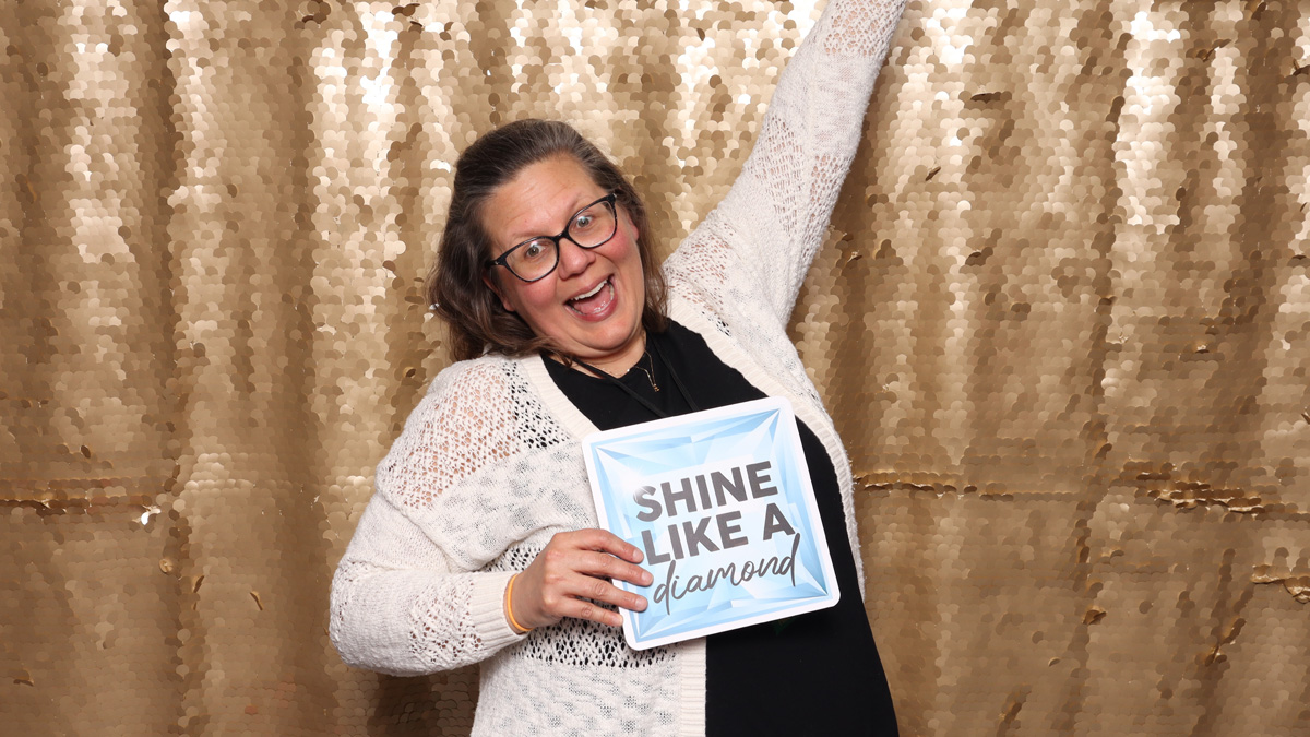 A white woman smiling with her arm in the air, standing in front of a gold, sparkly backdrop. She is holding a sign that reads, "Shine like a diamond."