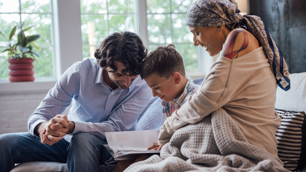 A mother and father read a book with their son. The mother has a headscarf and her shirt reveals the top part of a pink ribbon tattoo on her shoulder.