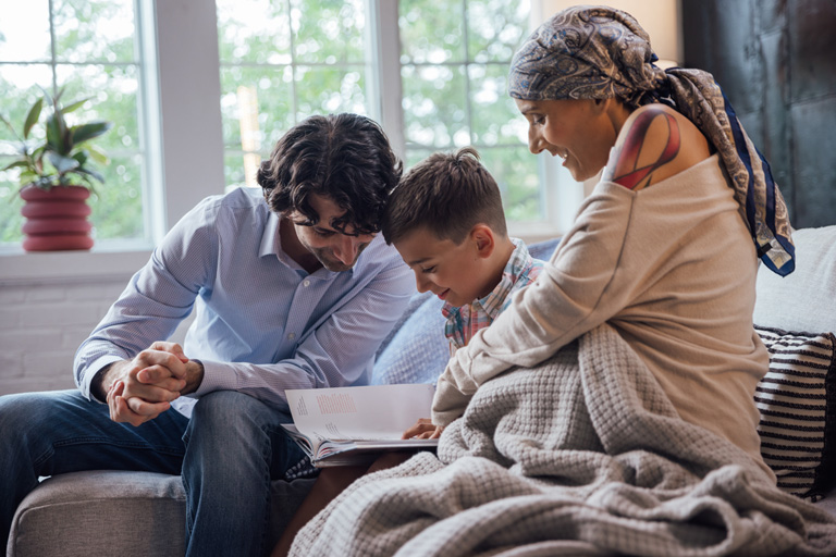A mother and father read a book with their son. The mother has a headscarf and her shirt reveals the top part of a pink ribbon tattoo on her shoulder.