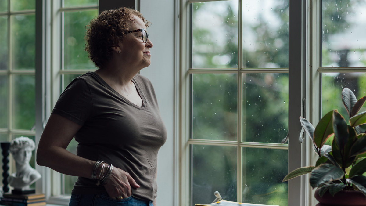 A white woman looking out the window with her hand in her pocket.
