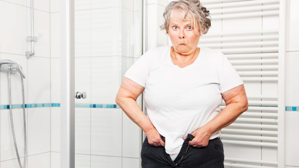 An older woman looks surprised as she tries to button her pants but cannot
