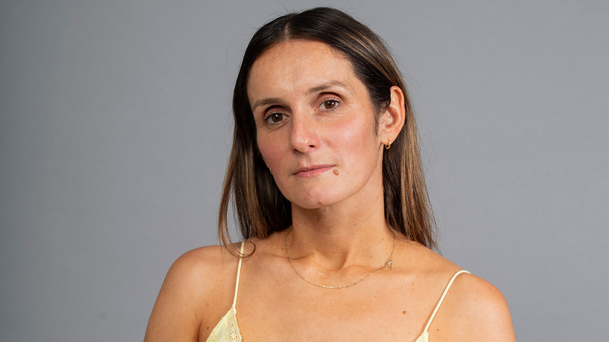 A white woman with a stoic look, standing in front of a grey backdrop