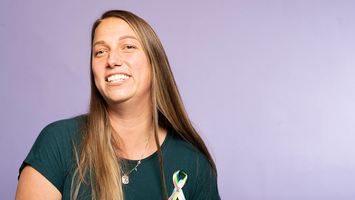 A white woman smiling in front of a purple background, wearing a ribbon with the MBC colors