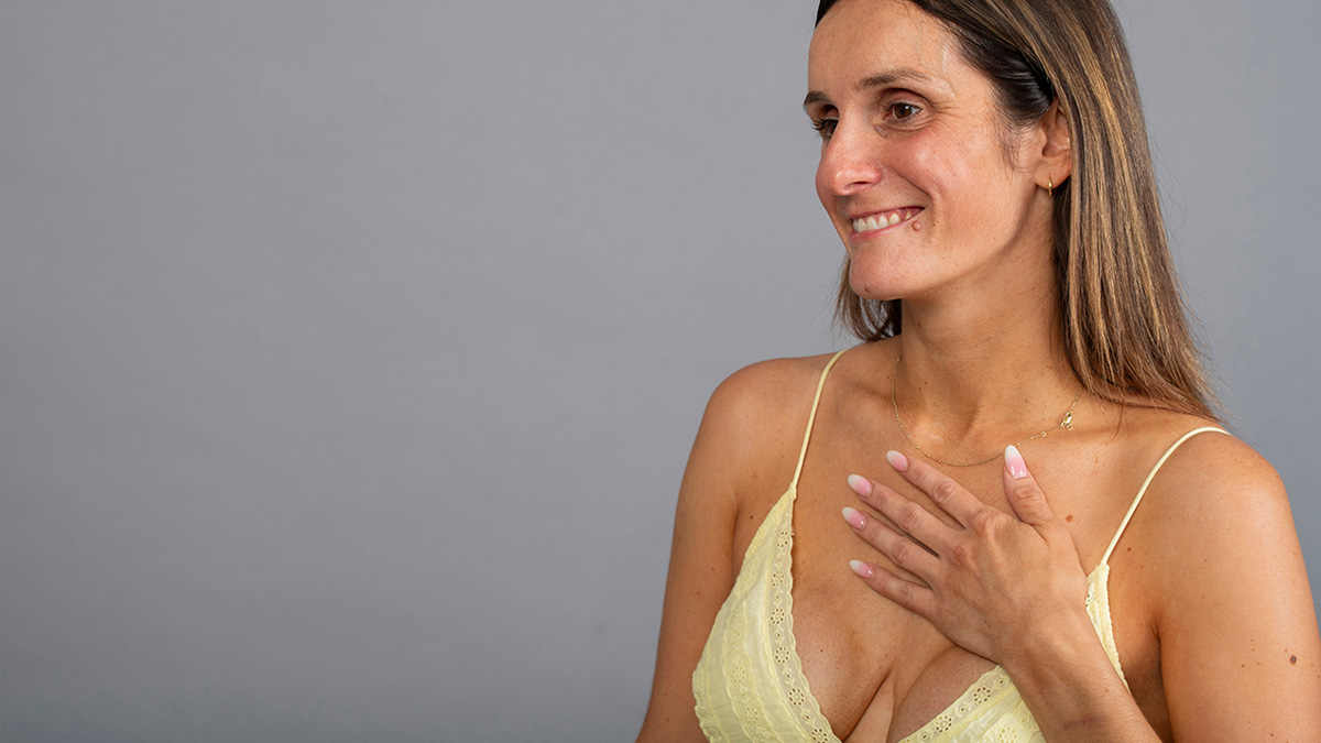 A white woman with a scar on her breast, holding her hand to her heart.