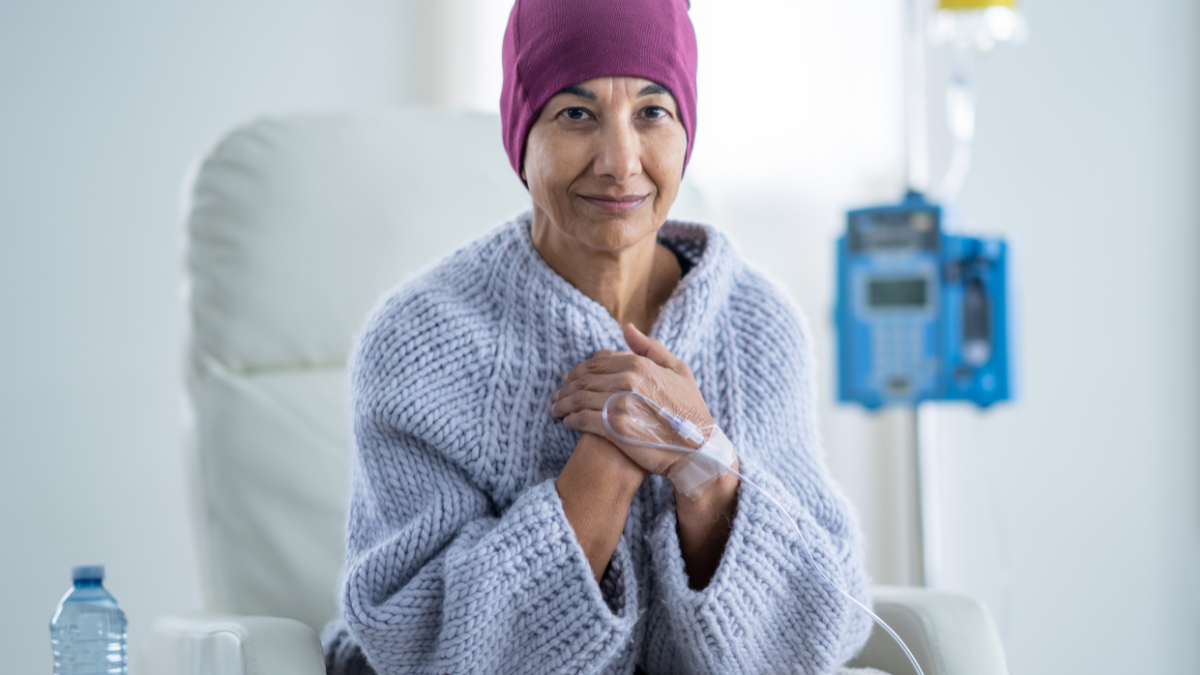 A woman sitting up in her treatment chair with an IV in her hand and a cap on her head