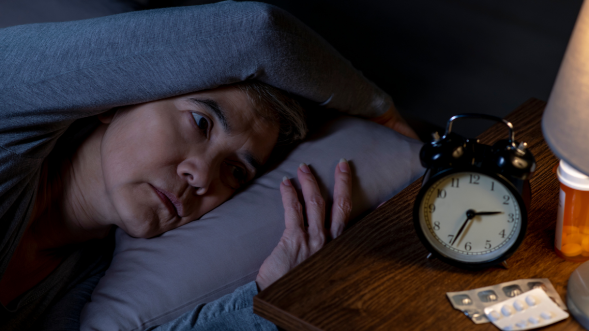 An older Asian woman laying awake in bed, looking at her clock with displays the time of 2:34 AM