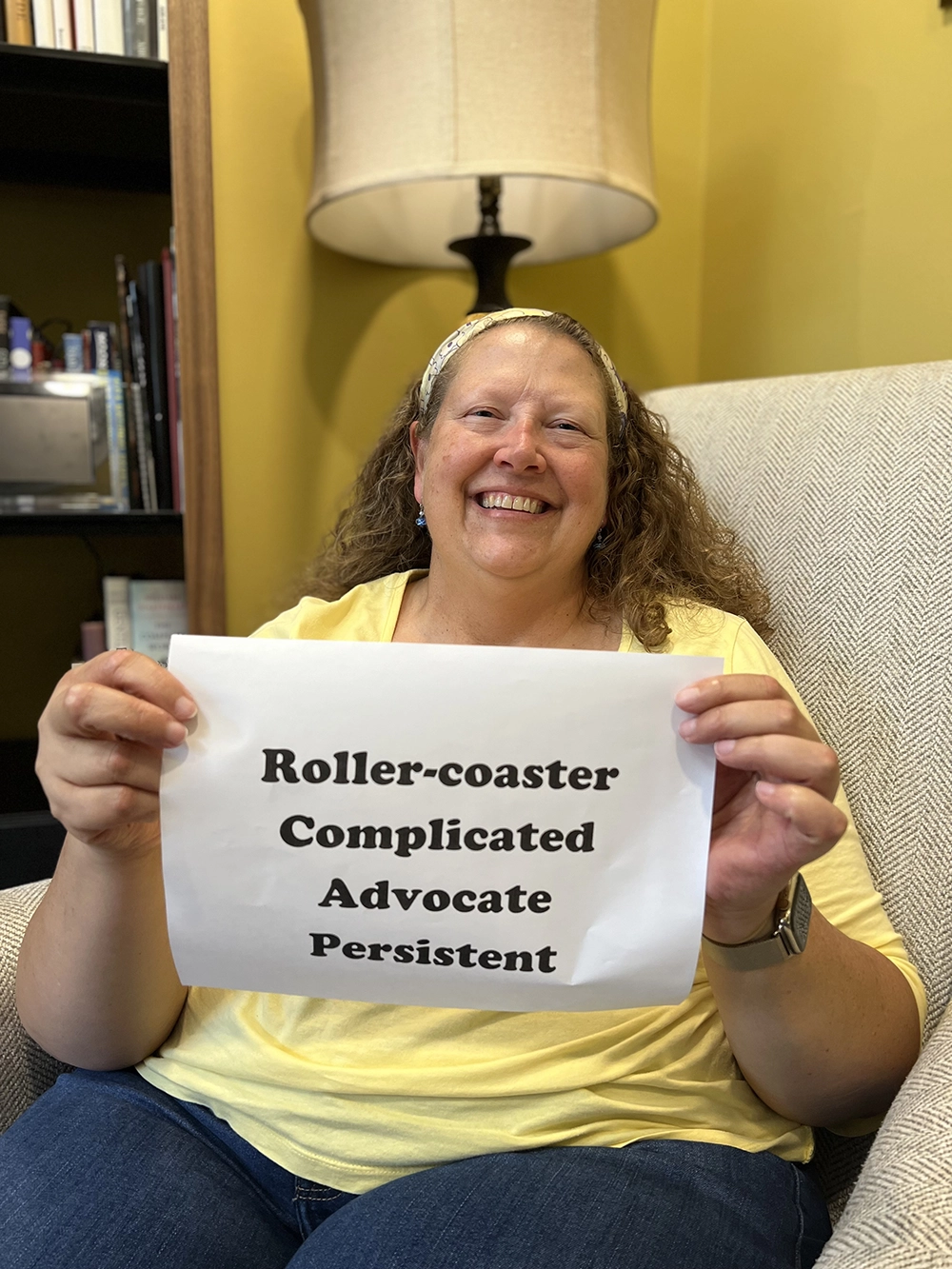 White woman with a small sign with the words Roller-coaster, Complicated, Advocate, and Persistent printed on it