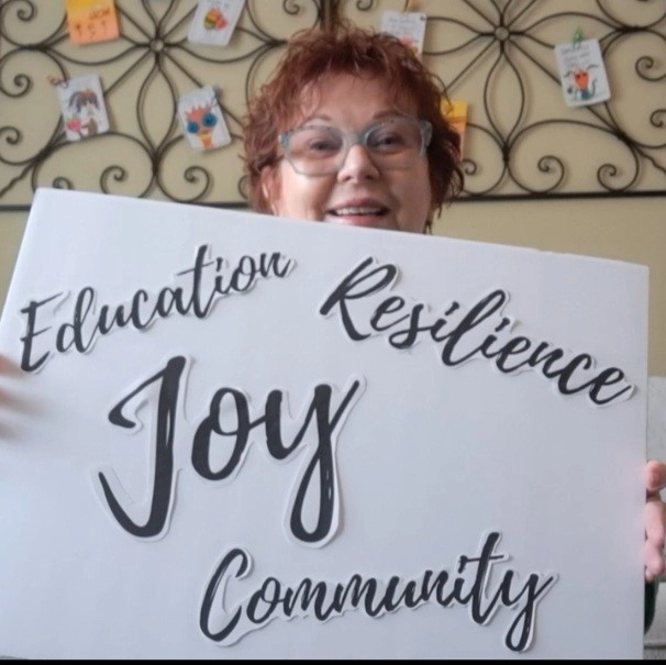 White woman holding white sign with the words Education, Resilience, Joy, and Community written on it in script