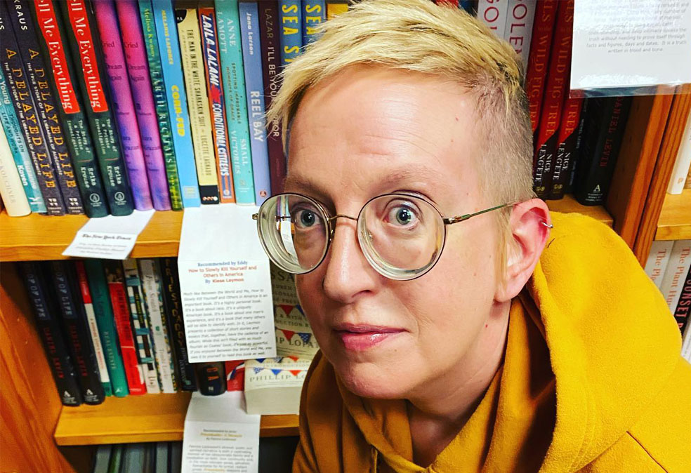 A young woman with thick glasses and a bright yellow hoodie in a bookstore