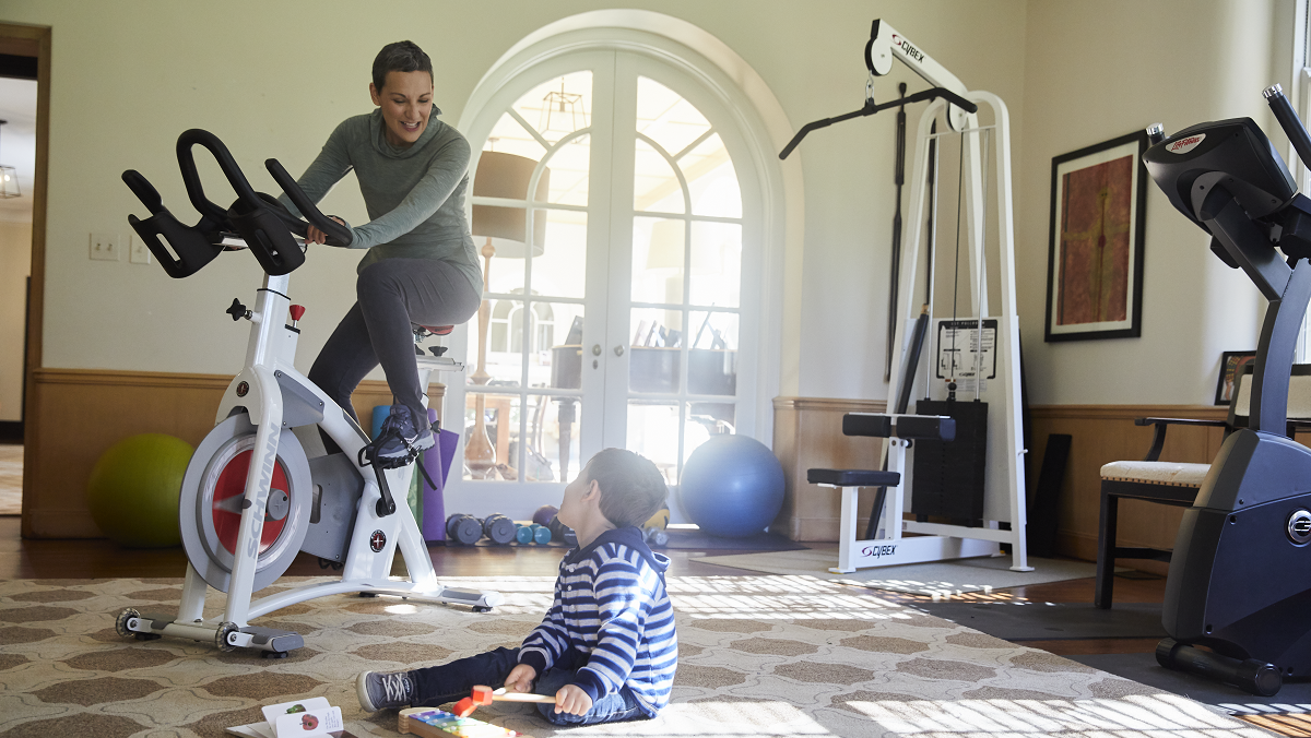 Woman on stationary bike looking at her young son on the floor next to her