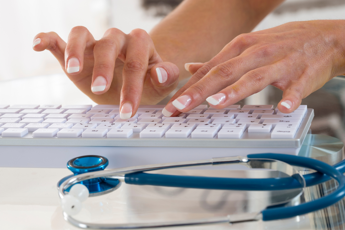 Close up of manicured hands typing on a keyboard, with a stethoscope lying on the table