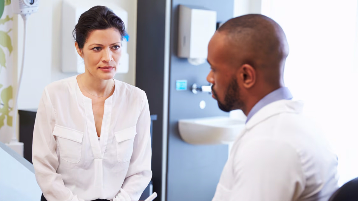 A white female patient in a doctor's office talking to a Black male doctor.