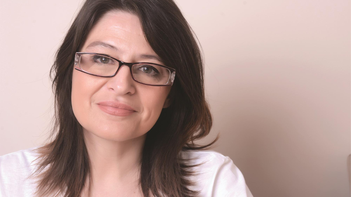 A white woman with rectangular glasses sitting against a pale pink background