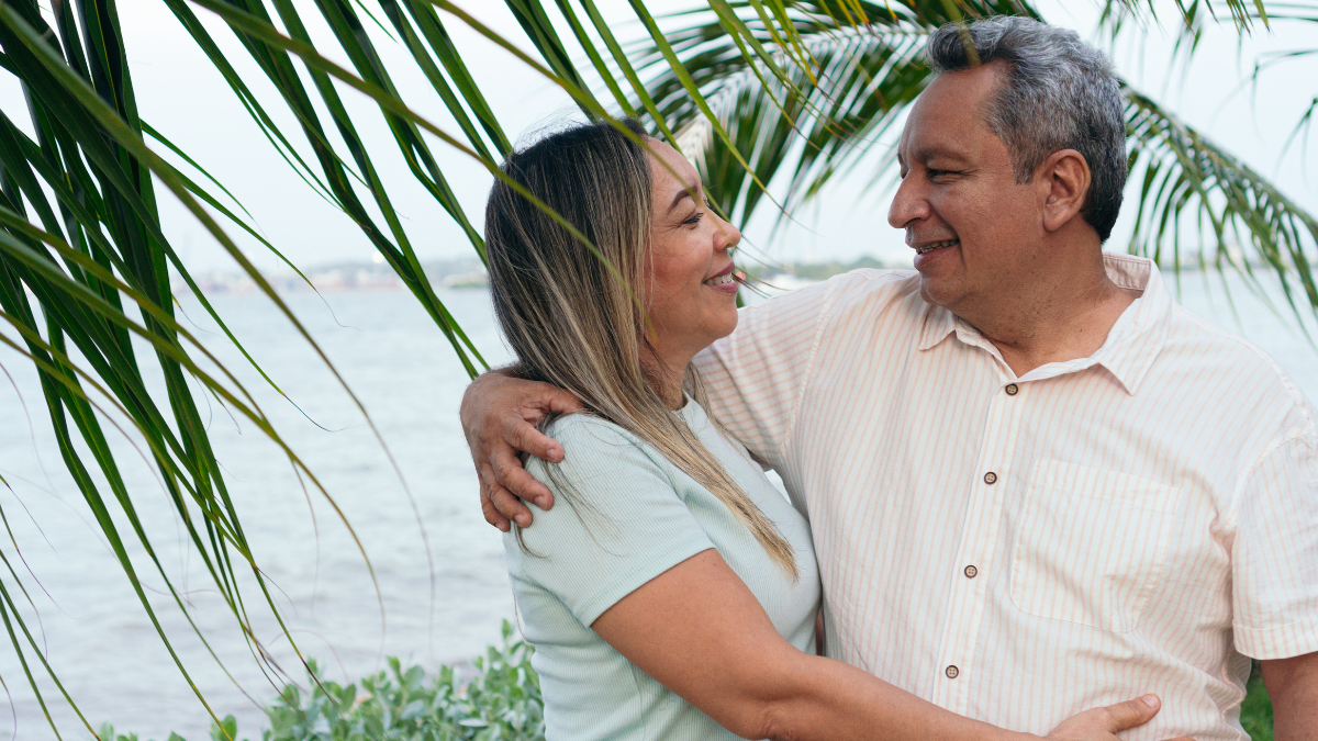 A Latino couple with grey hair embrace and look lovingly at each other as they stand beneath a palm tree.