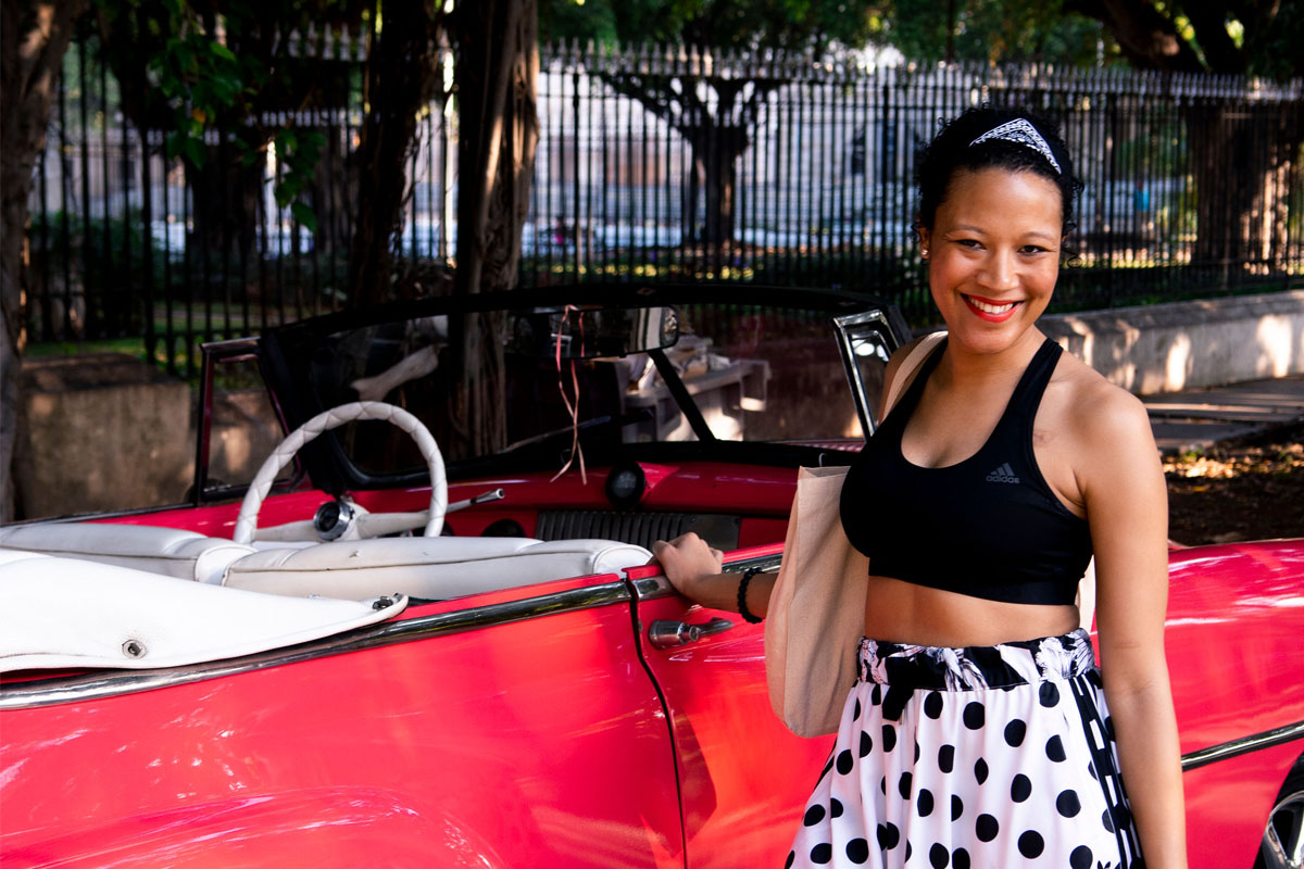 A young Black woman stands by a vintage car in Cuba