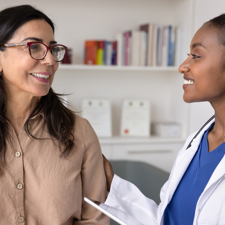 An older woman with glasses and her female, Black doctor smile at each other in a doctor's office.