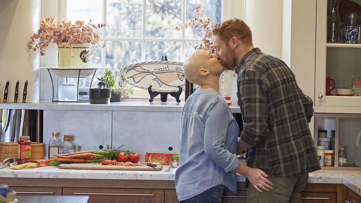 A white heterosexual couple kiss in the kitchen. The woman has a shaved head.