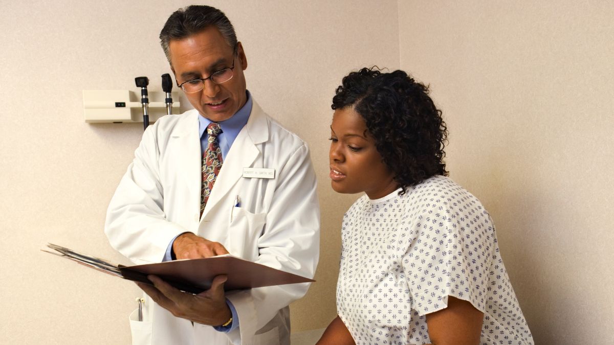 A male doctor shows a young Black woman dressed in a hospital gown something in a medical record