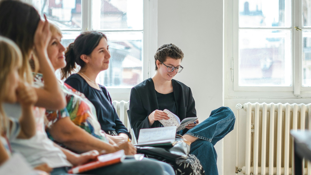 White women sitting in chairs with notebooks looking at someone.