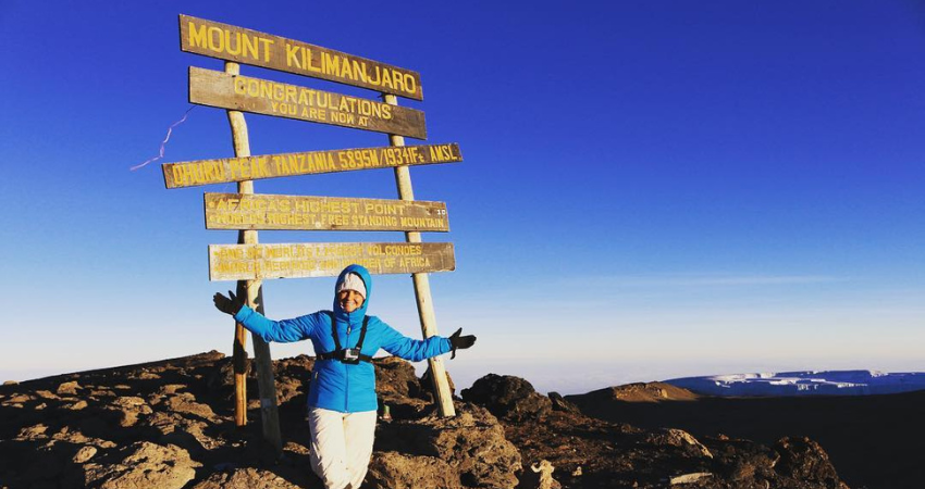 Gillian posing at the top of Mount Kilimanjaro with "Congratualtions" sign behind her