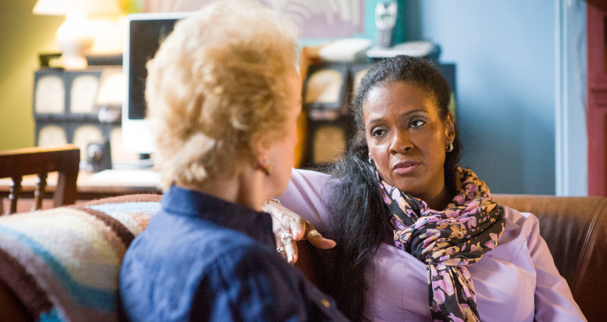 An older Black woman and an older white woman sit on a couch, facing each other and talking seriously
