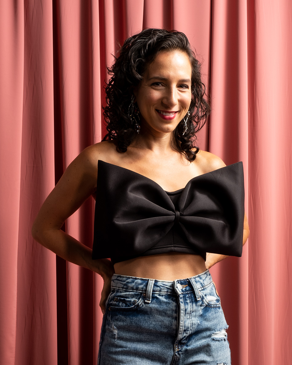 Abby smiling in a top with an overly large black bow and blue jean pants. Photo by Ken Cox and Andrew Scherer from Kronus Studio
