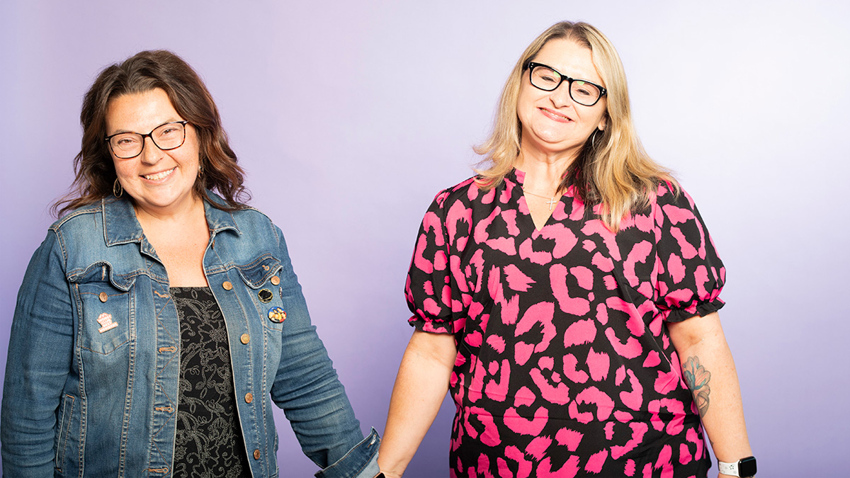 Two white women holding hands and smiling in front of a purple backdrop