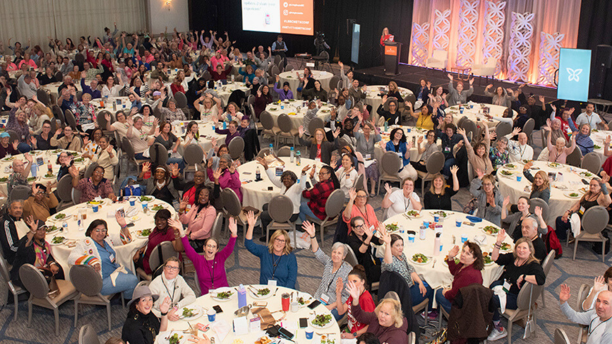 Hundreds of conference attendees sitting at their tables, waving at the camera, at the 2025 Mets Conference