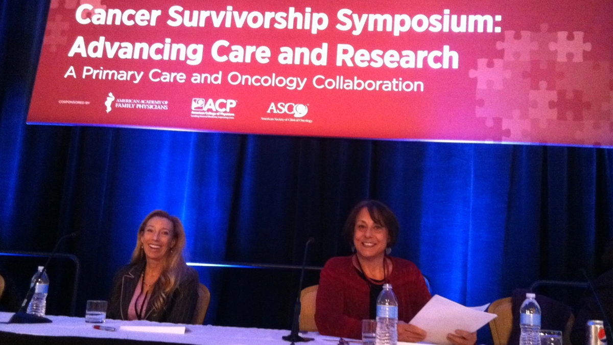 Diane at the ASCO Survivorship Symposium in 2016, sitting at a long table under a large sign that reads, "Cancer Survivorship Symposium: Advancing Care and Research"