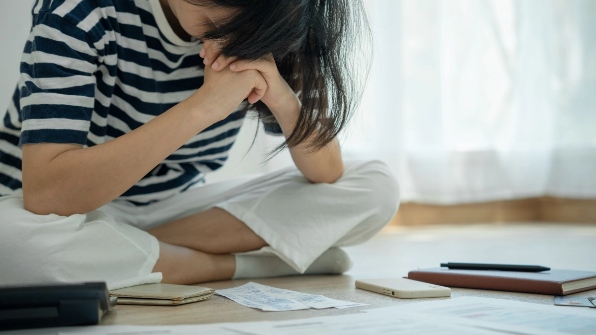 A distressed women with black hair wearing a black and white striped shirt and white pants sits on the floor amid bils.