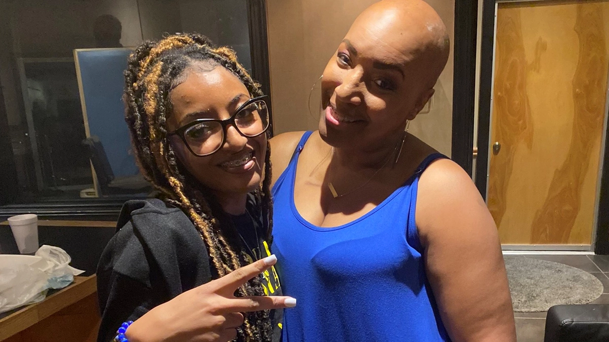 Bald Keneen in blue tank dress with Kalista, who has long braided hair, glasses and makes a 'V' for victory with her index and middle fingers.