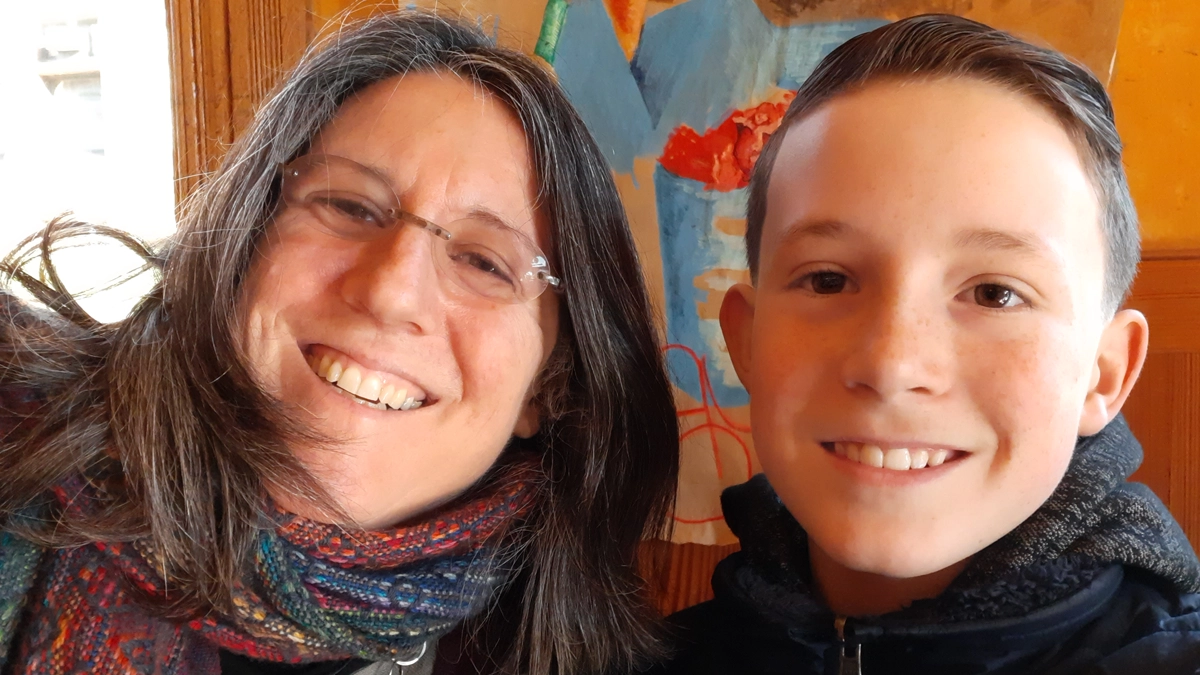 Sheri, gray and brown long hair wears glasses and smiles beside her sone, with short brown hair and freckles.