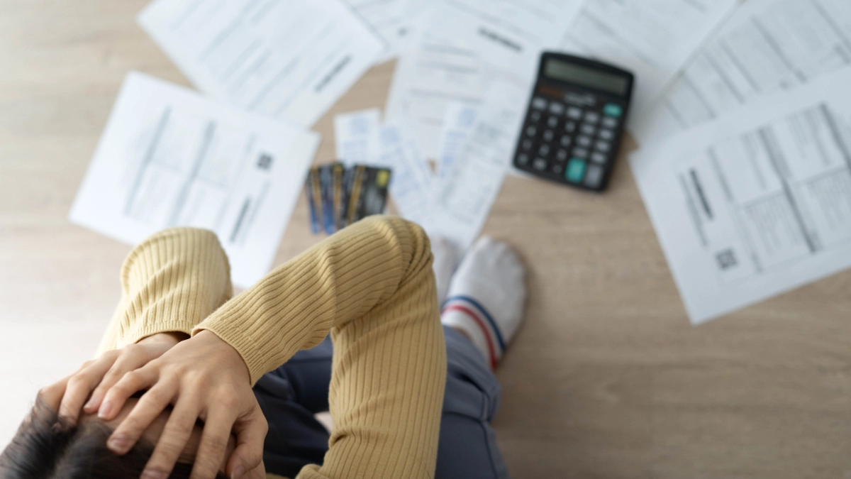 Person with hands covering face sits on floor that is covered with papers and a calculator.