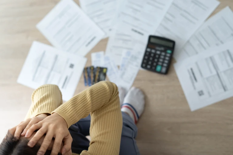 Person with hands covering face sits on floor that is covered with papers and a calculator.