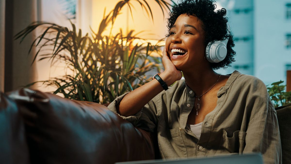A young Black woman sits on a couch, listening to headphones with a large smile