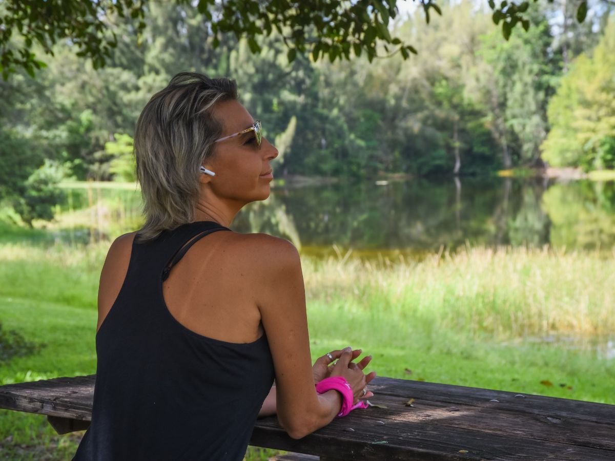 Patricia, a Latina woman, sits at a picnic table under a tree. She is wearing workout clothes and has earbuds in.