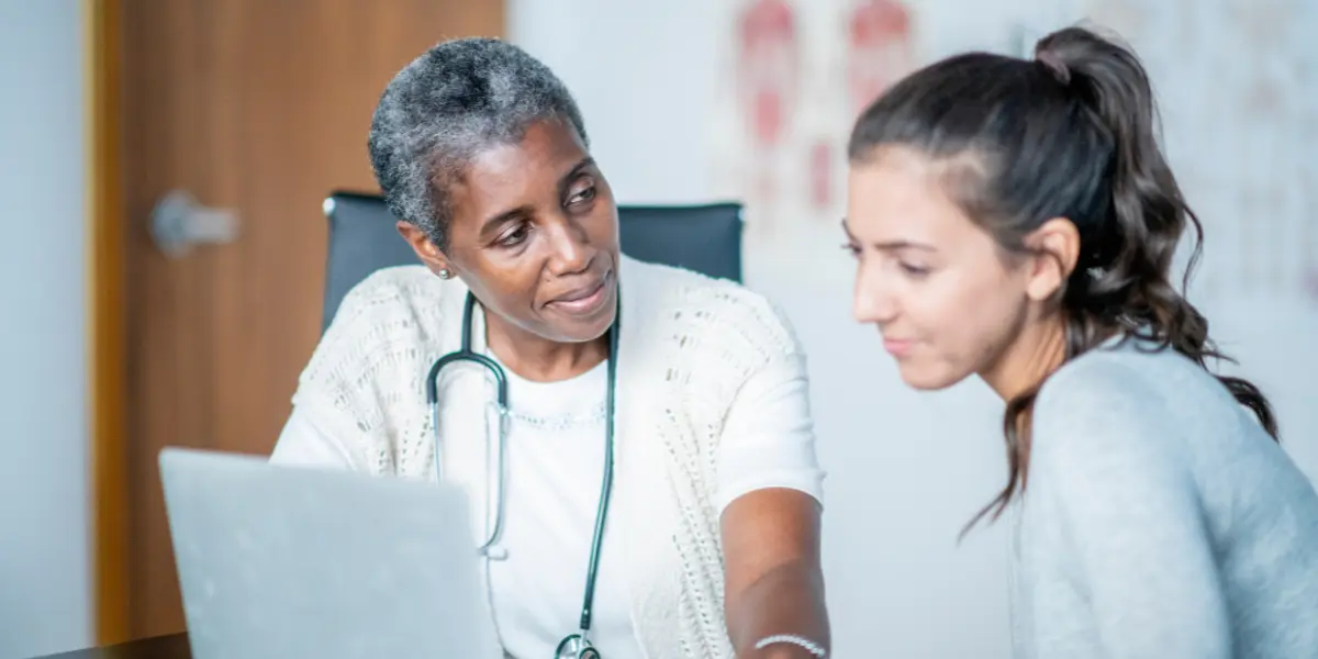 An older Black woman with a stethoscope around her neck talks to a young white woman about something on a laptop in front of them both