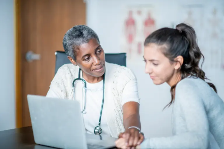 An older Black woman with a stethoscope around her neck talks to a young white woman about something on a laptop in front of them both