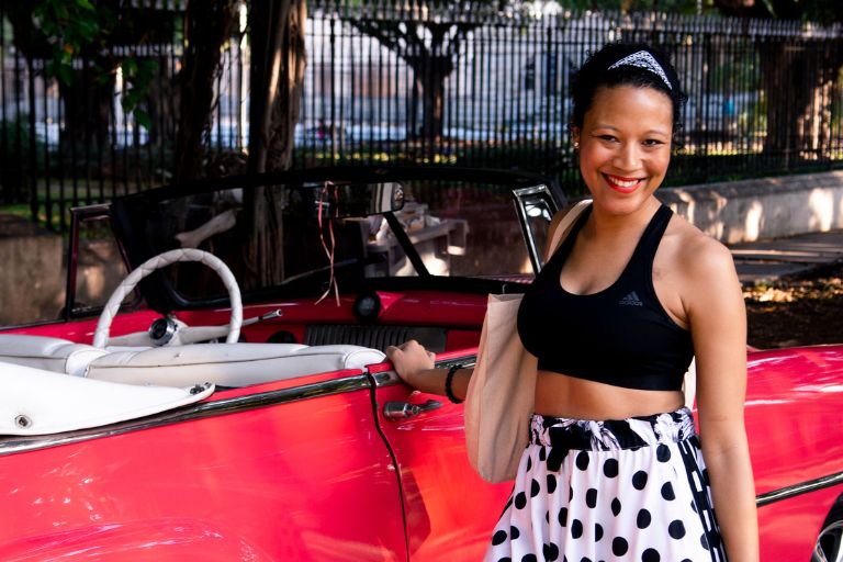 A young Black woman stands by a vintage car in Cuba